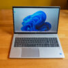 Silver laptop on wooden table displaying Windows 11 desktop with blue swirl wallpaper.