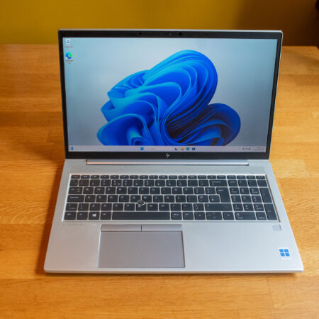 Silver laptop on wooden table displaying Windows 11 desktop with blue swirl wallpaper.