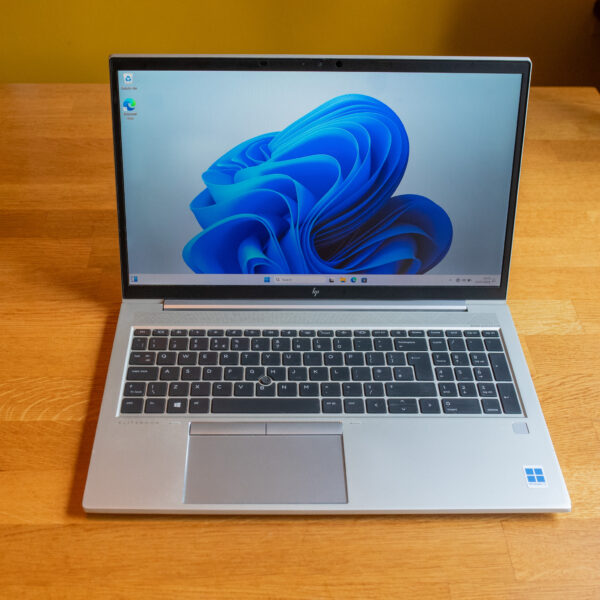 Silver laptop on wooden table displaying Windows 11 desktop with blue swirl wallpaper.