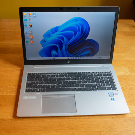 Laptop with Windows 11 desktop, placed on wooden table, displaying blue swirl wallpaper.